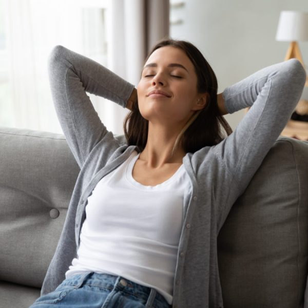 Relaxed serene pretty young woman feel fatigue lounge on comfortable sofa hands behind head rest at home, happy calm lady dream enjoy wellbeing breathing fresh air in cozy home modern living room